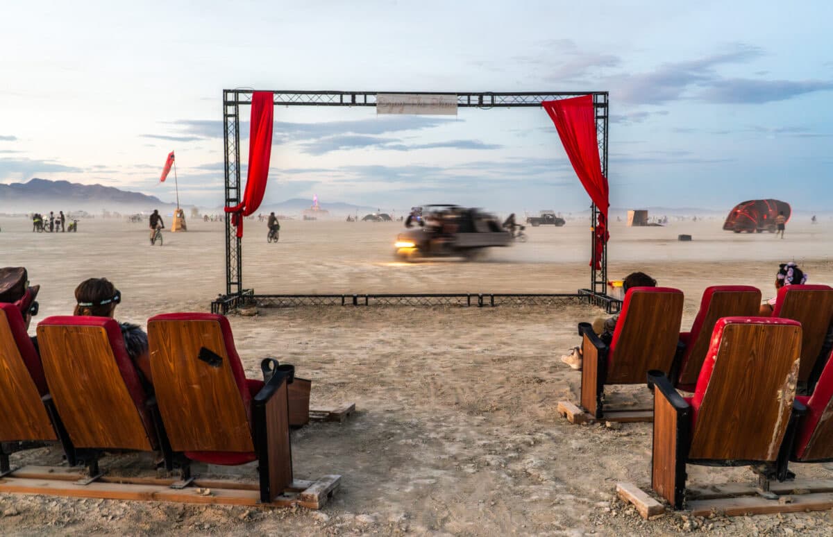 Salvaged cinema seats facing an open proscenium with red curtains while an art car blurs past across the playa.
