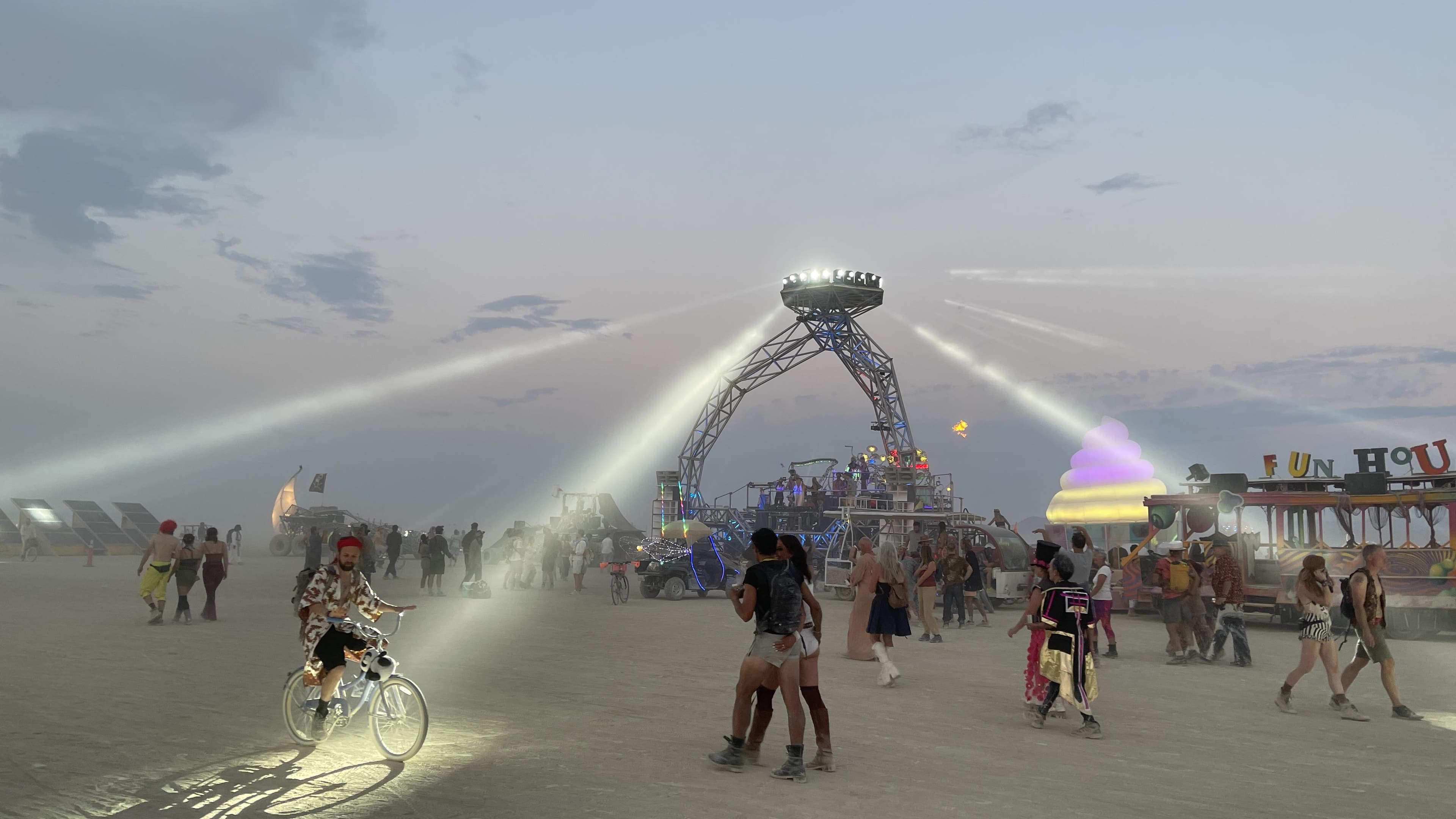 A rider on a lit bike throws a beam of light across the dust at dusk while everyone else fades to a gray shape.