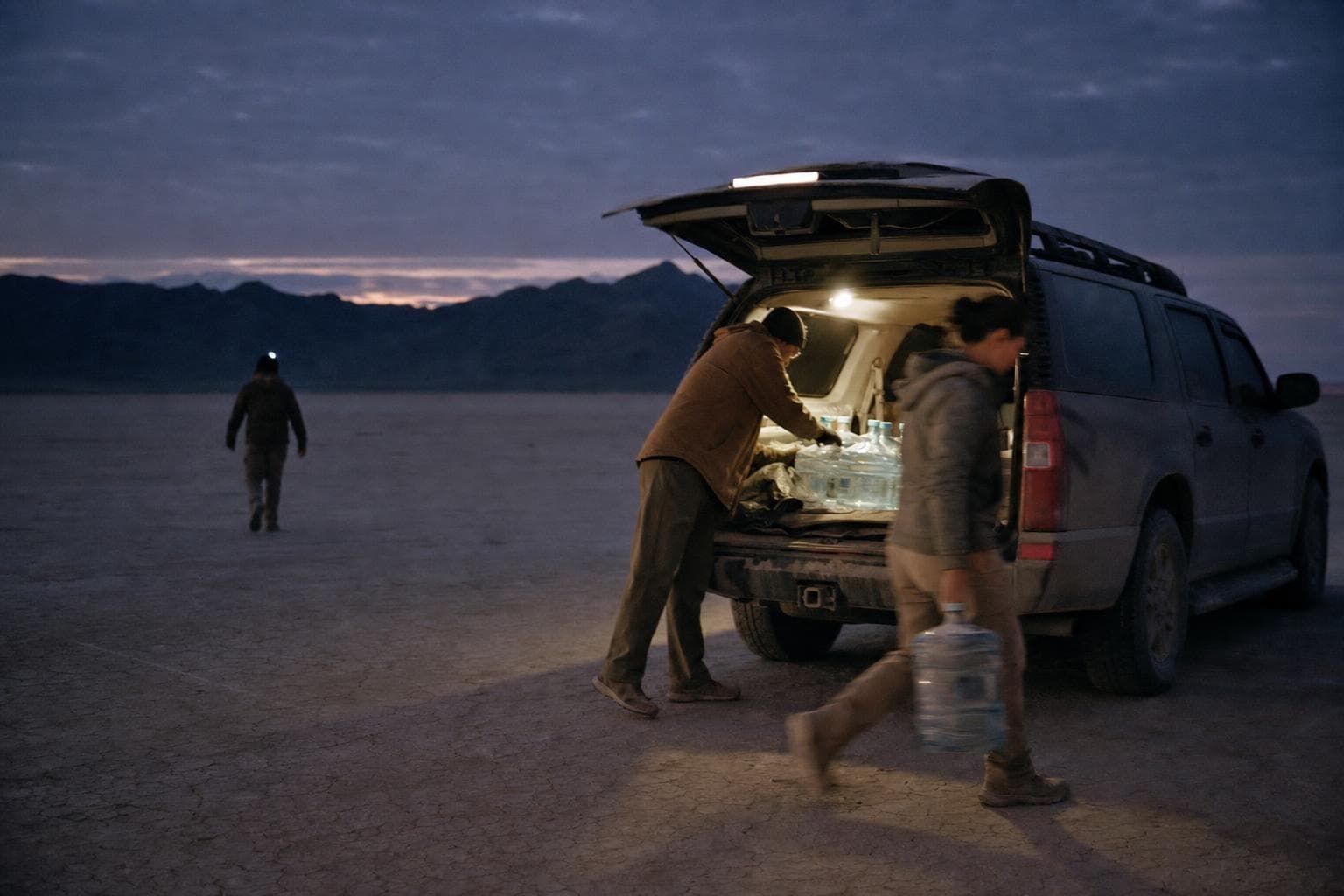 A dome light glowing in an open tailgate at dusk while someone lifts water jugs and a headlamp walks off into the flat.