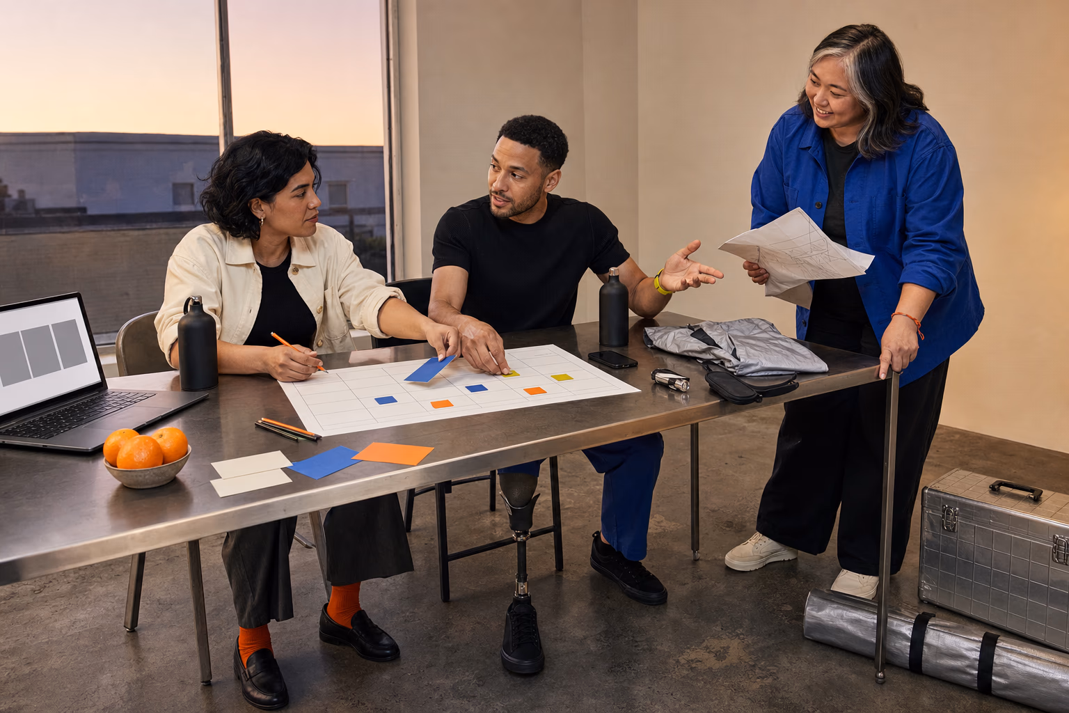 Three friends building a shared trip plan with a paper grid, gear tokens, textiles, and notes around a studio table